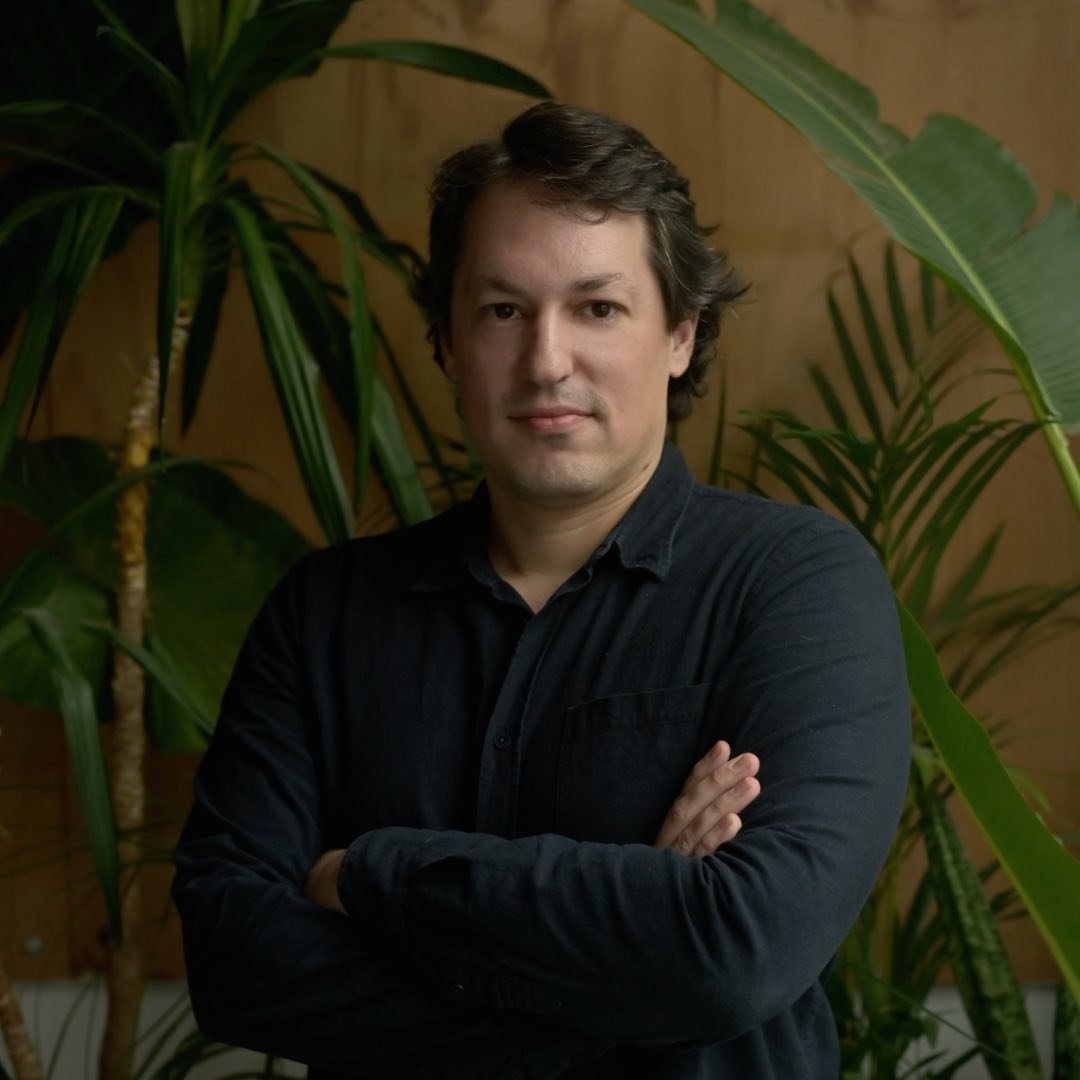 Shahar in green, arms folded with a palm tree behind him.